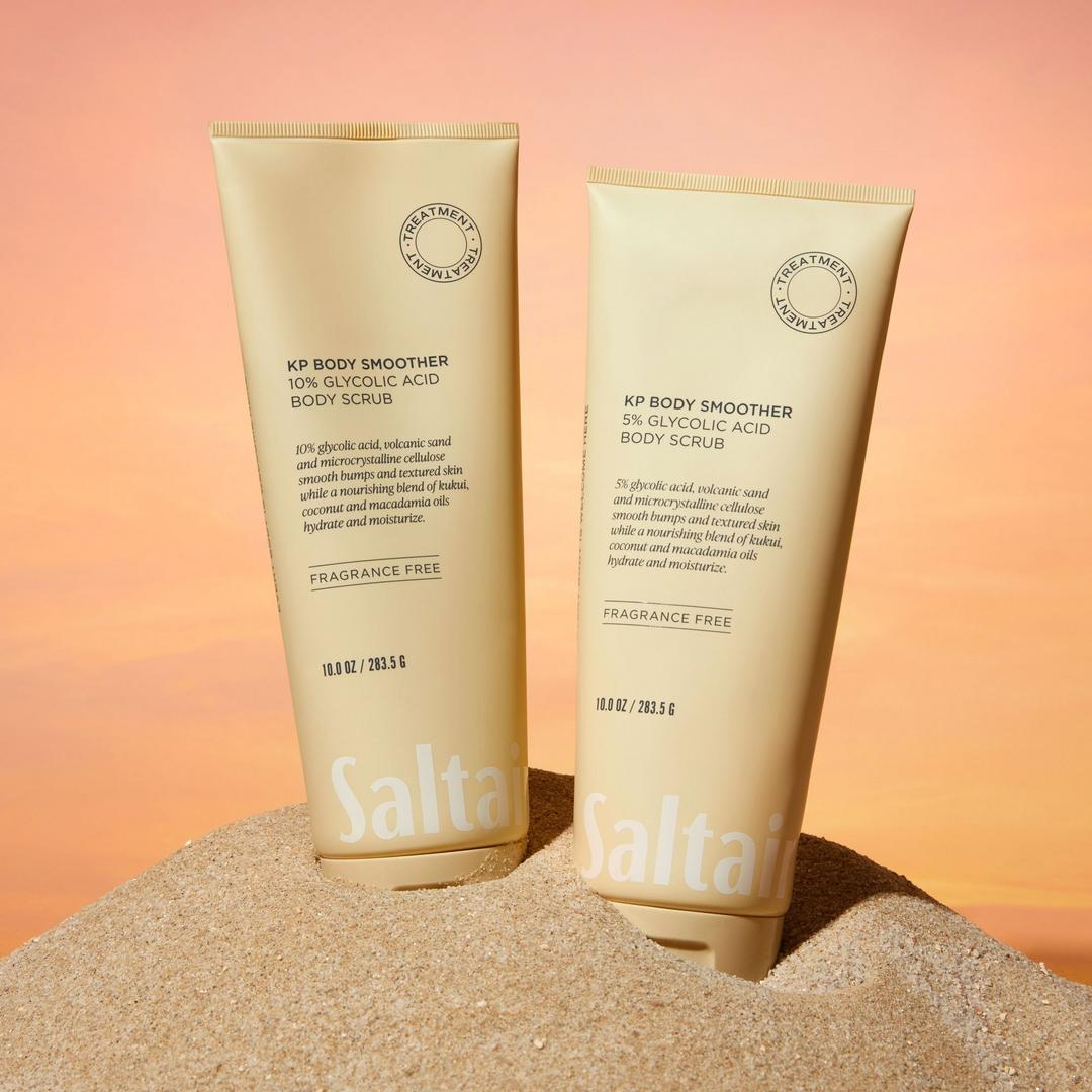 Saltair KP Body Smoother 5 percent and 10 percent Glycolic Acid Body Scrubs displayed upright on sand against a warm sunset background.