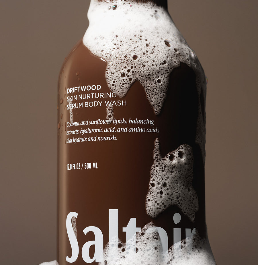 Saltair Driftwood Serum Body Wash bottle covered in foam on a brown background.